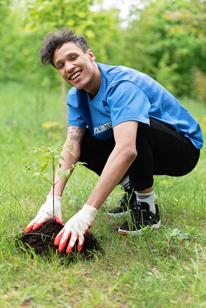 A smiling volunteer plants a young tree outdoors, wearing gloves, in a spring garden.