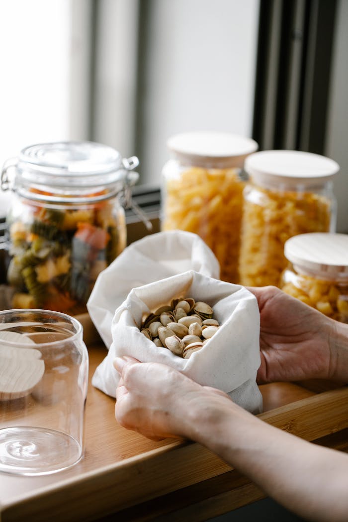 Close-up of hands holding pistachios in a bag next to jars of dried food on a wooden table.